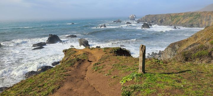 A path ending in a rocky coastline with waves crashing on rocks below the end of the path.