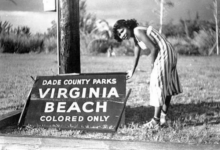 A Black woman in beachware holds up a sign that says, "Virginia Beach: Colored Only"