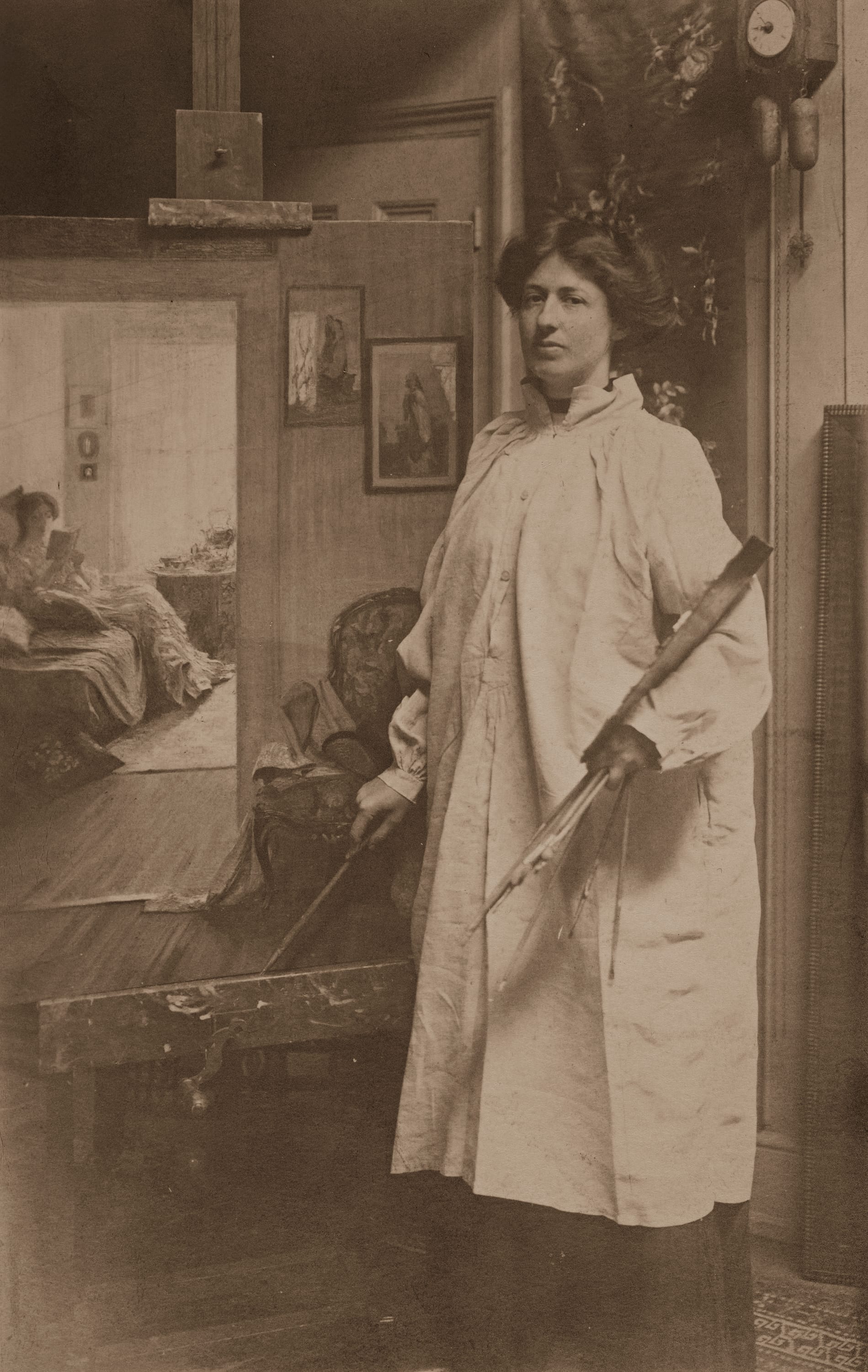 A woman wearing a paint smock stands in her studio holding brushes and a palette beside an easel displaying a painting of a seated figure reading.
