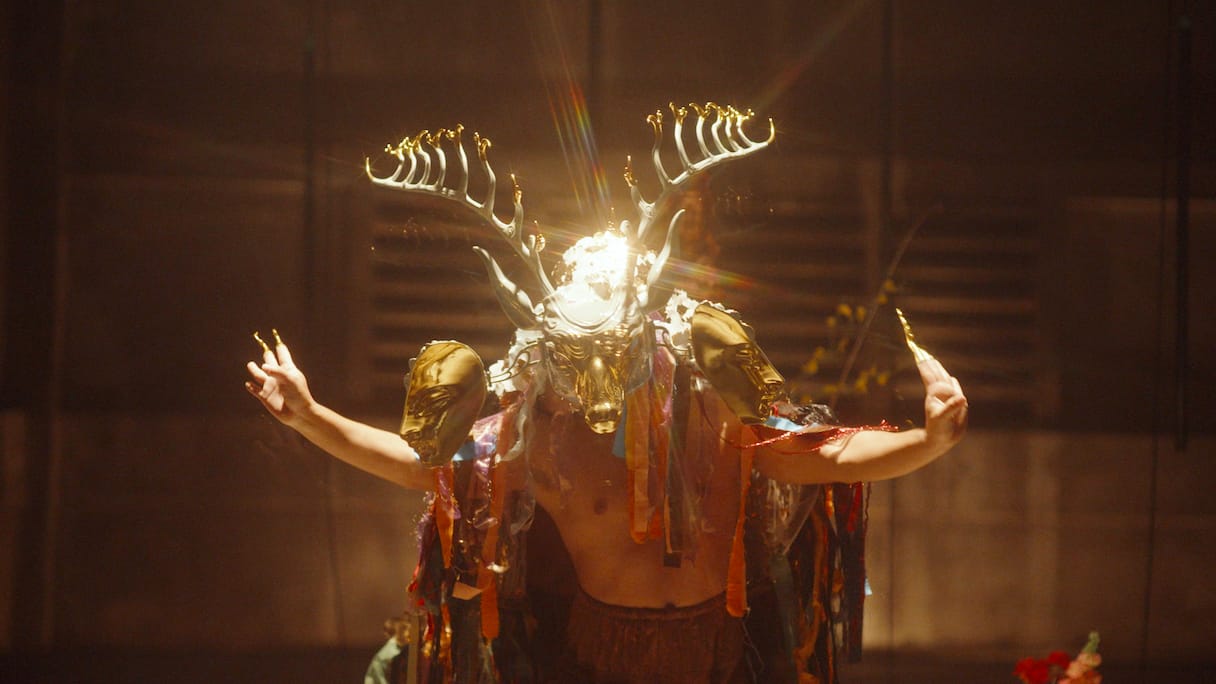 Performer wearing an elaborate gold deer mask with antlers and additional gold face pieces, surrounded by colorful ribbons, illuminated dramatically on a dim stage.