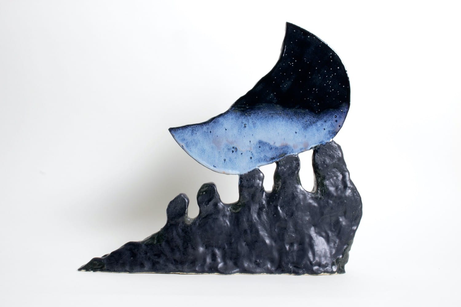 Small sculptural form resembling a crescent moon balanced atop a jagged, dark base, with a speckled blue surface against a white background.