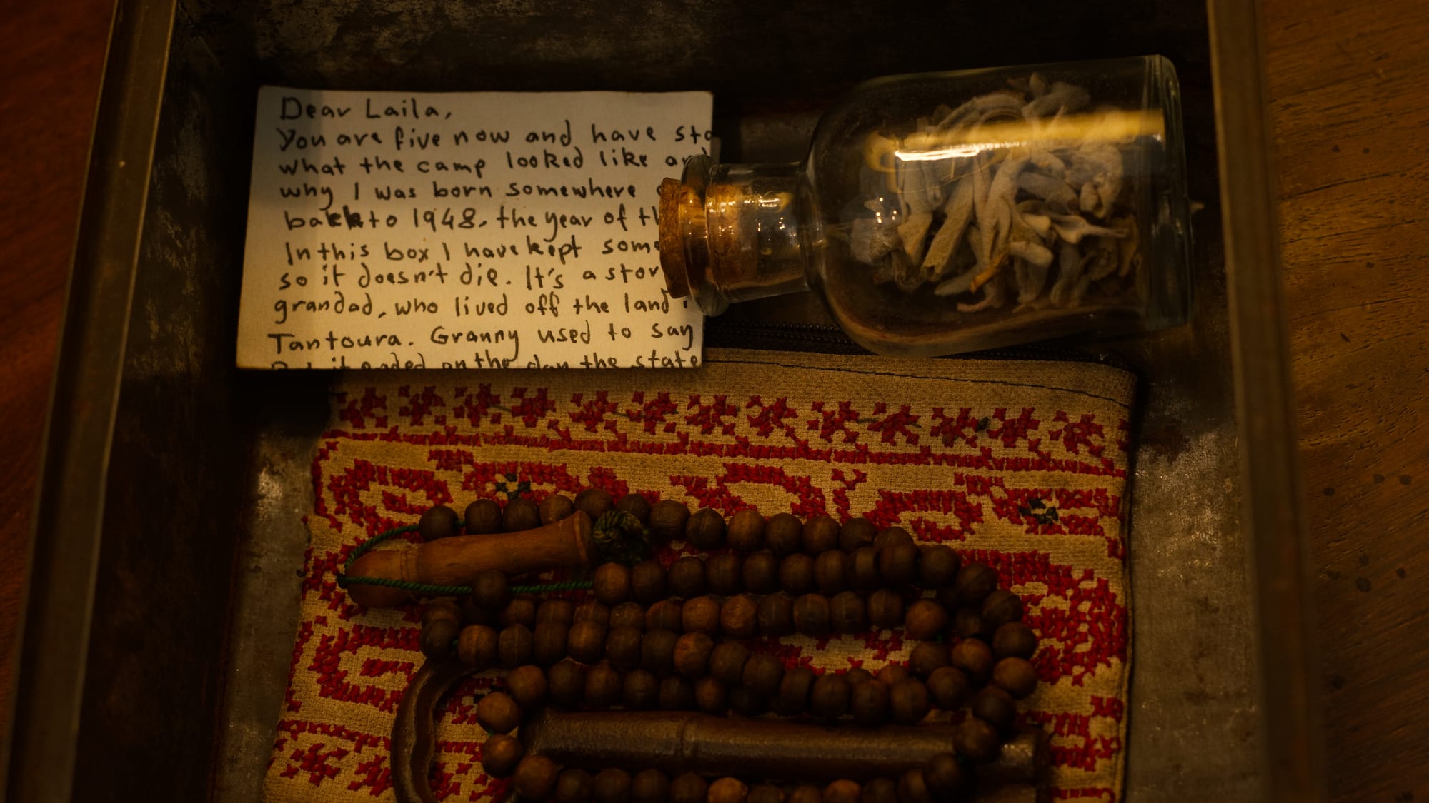 Close-up of a box containing a handwritten note, a small bottle of dried leaves, prayer beads, and embroidered fabric.