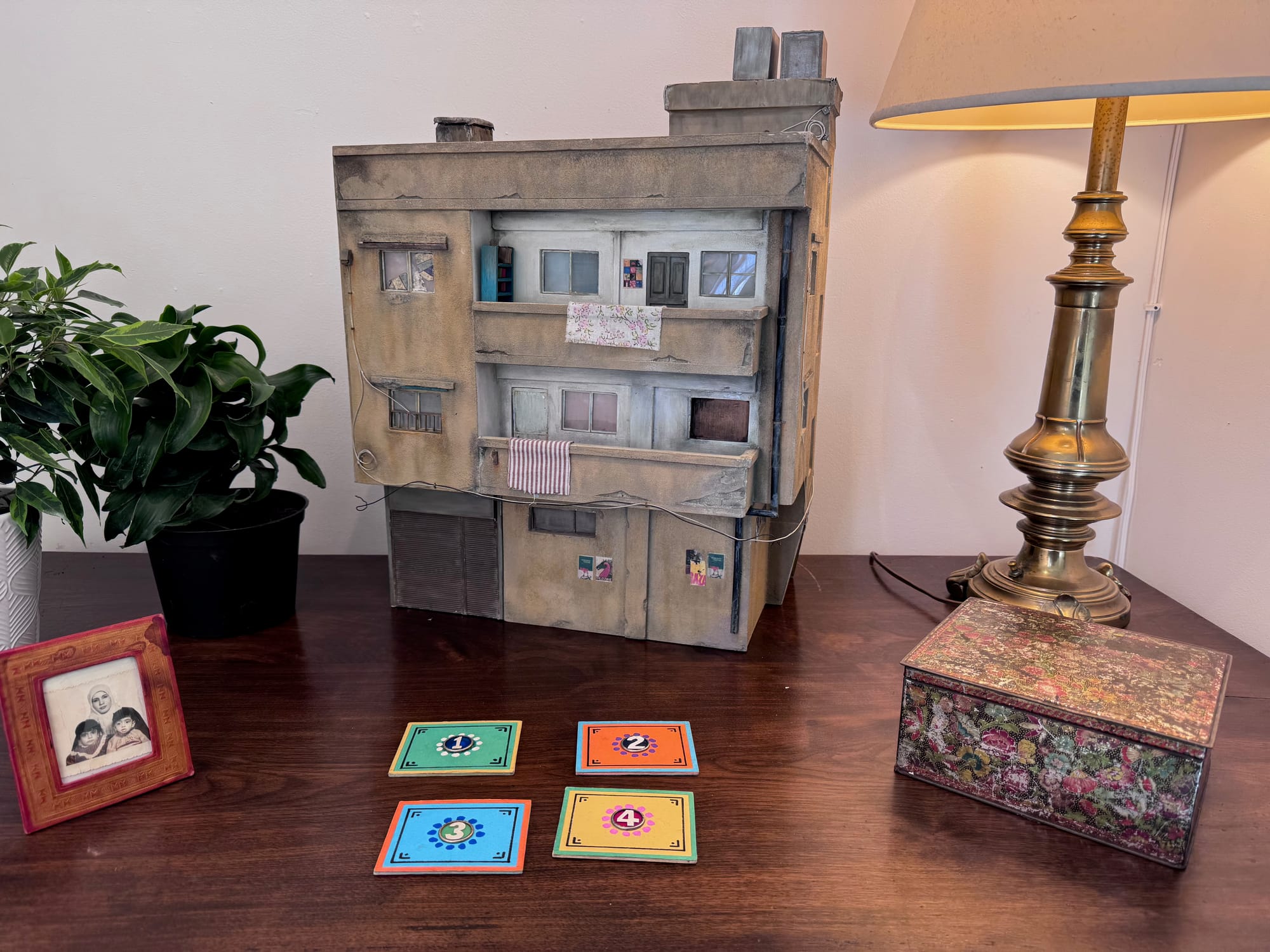Model of a multi-story building on a desk with numbered cards, a framed photo, a plant, a lamp, and a decorative box.
