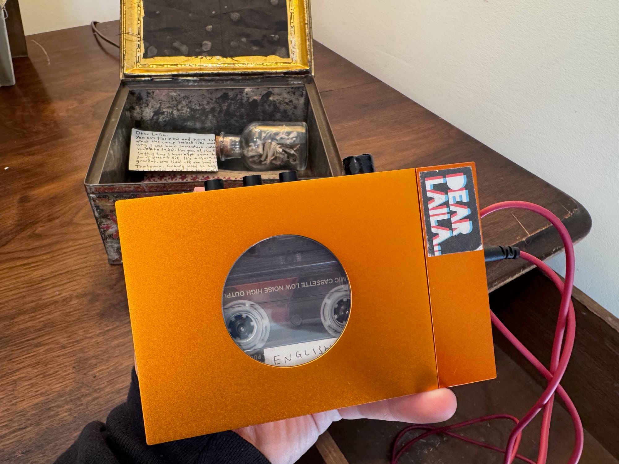 Hand holding an orange cassette player connected by a cable, with an open box containing a note and small bottle in the background.