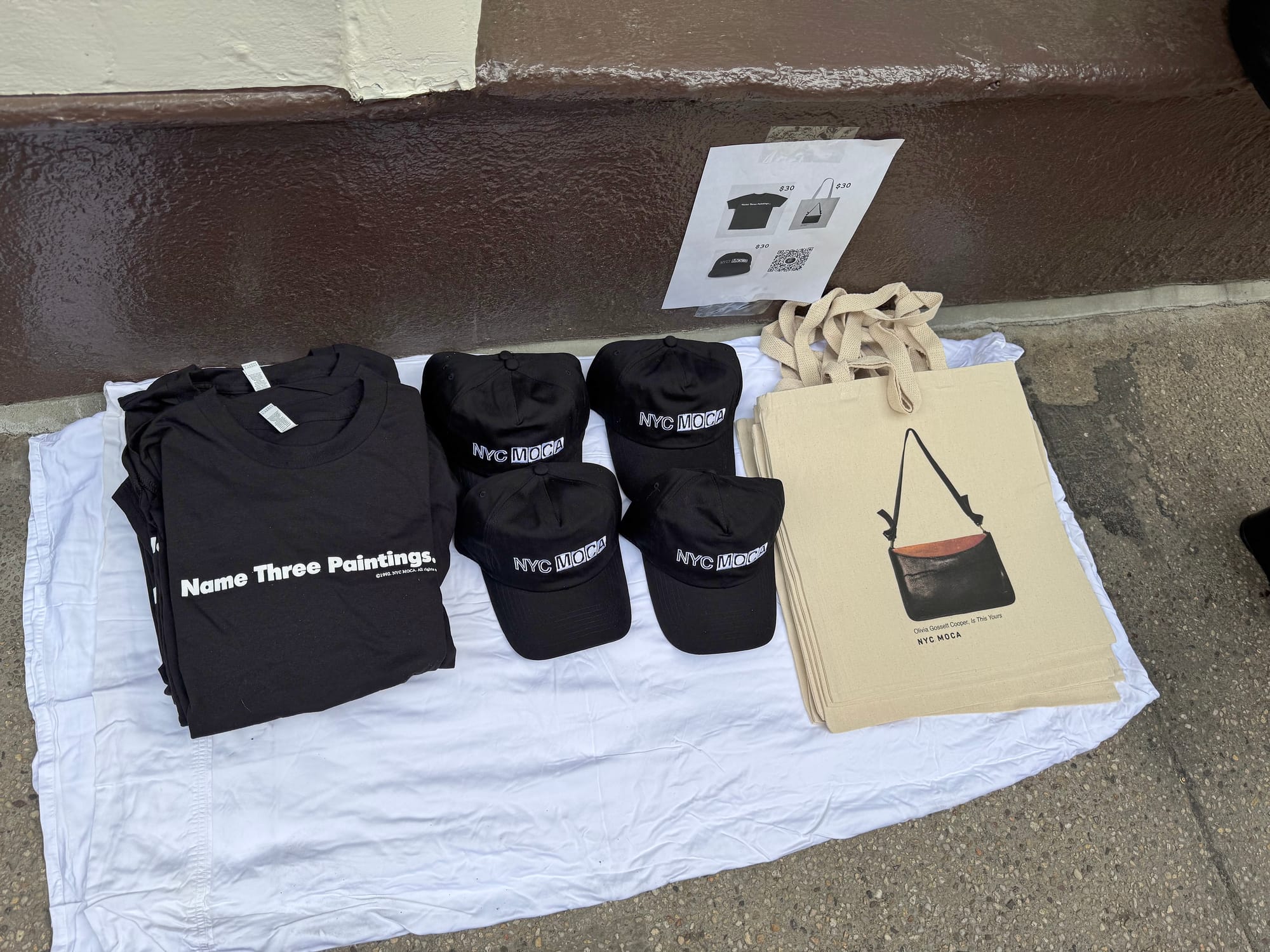 black shirts and hats with NYC MOCA branding are seen on a white towel, along with NYC MOCA-branded tote bags