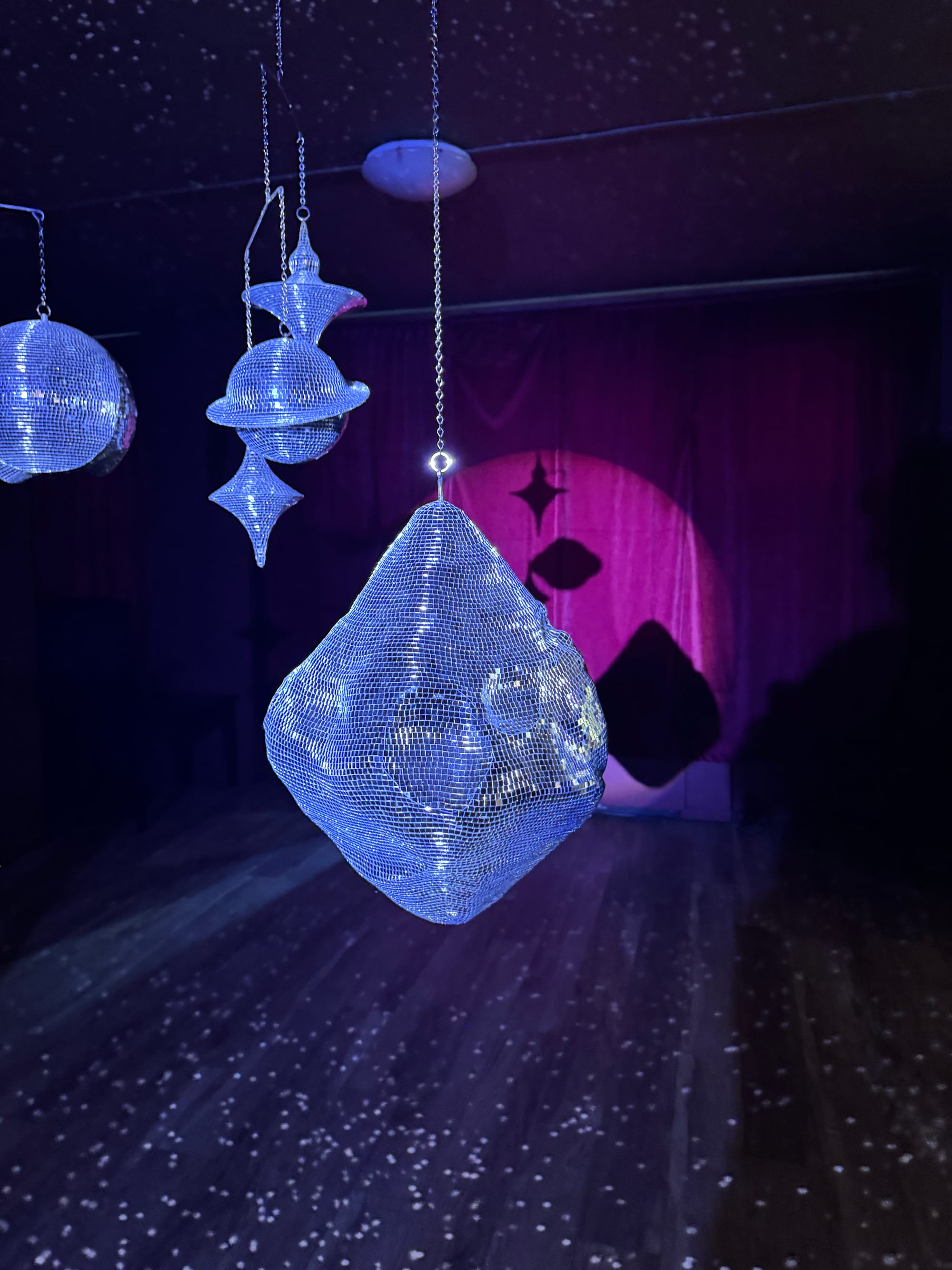 Mirrored, hanging sculptural forms reflect light in a dark gallery, casting shifting patterns against a pink-lit backdrop.