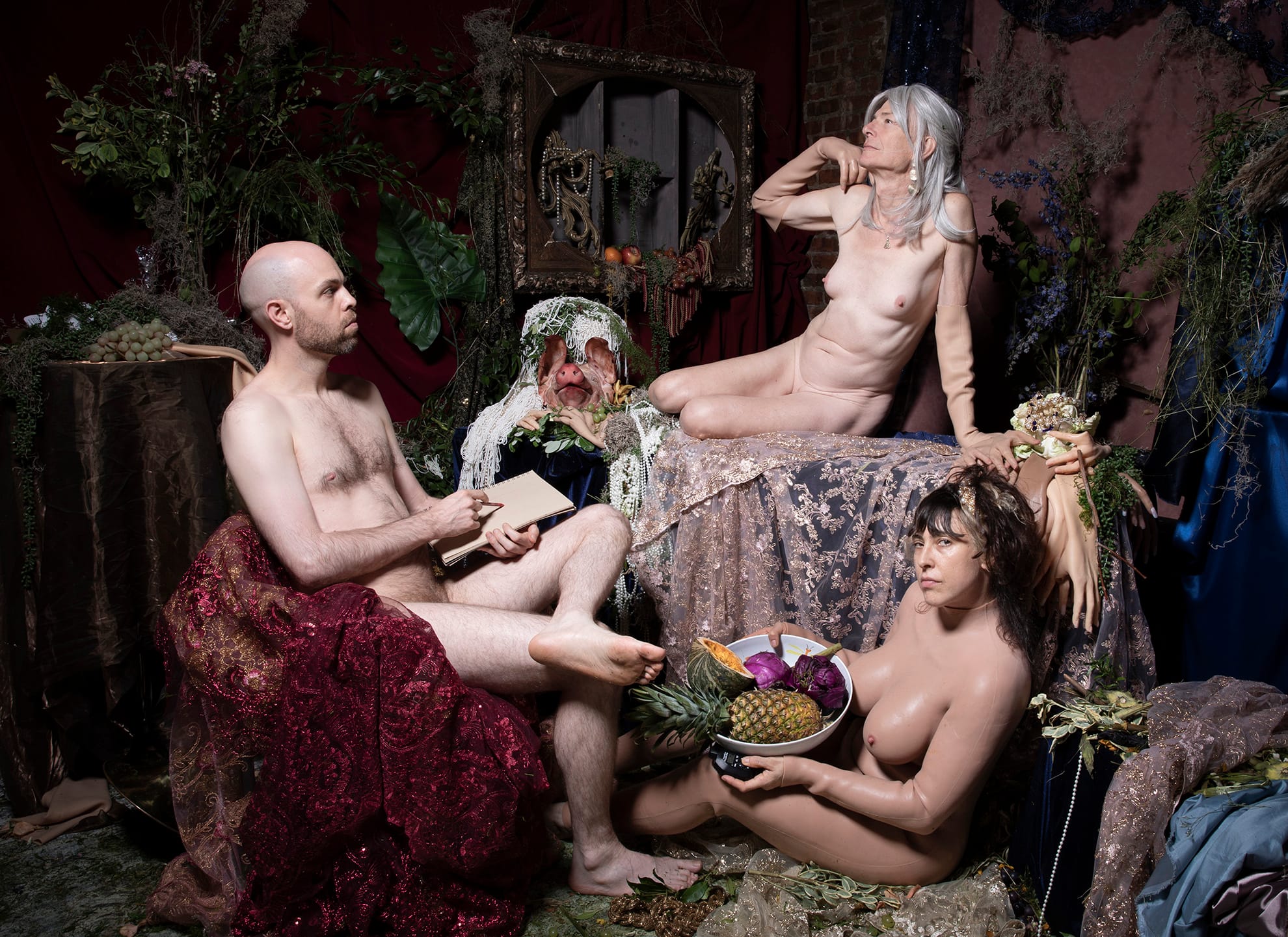 A staged scene of several nude figures amid drapery and fruit; a bald man at left sketches in a notebook, a silver-haired figure reclines at upper right, and a woman holds a bowl of fruit at lower right, with a veiled pig's head at center.