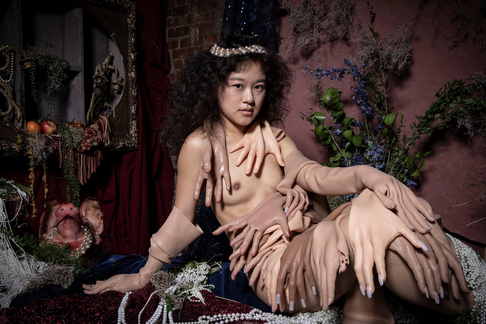A young nude woman in a pearl headpiece reclines covered in dozens of flesh-colored prosthetic gloves and hands, against velvet, flowers and a pig's head.