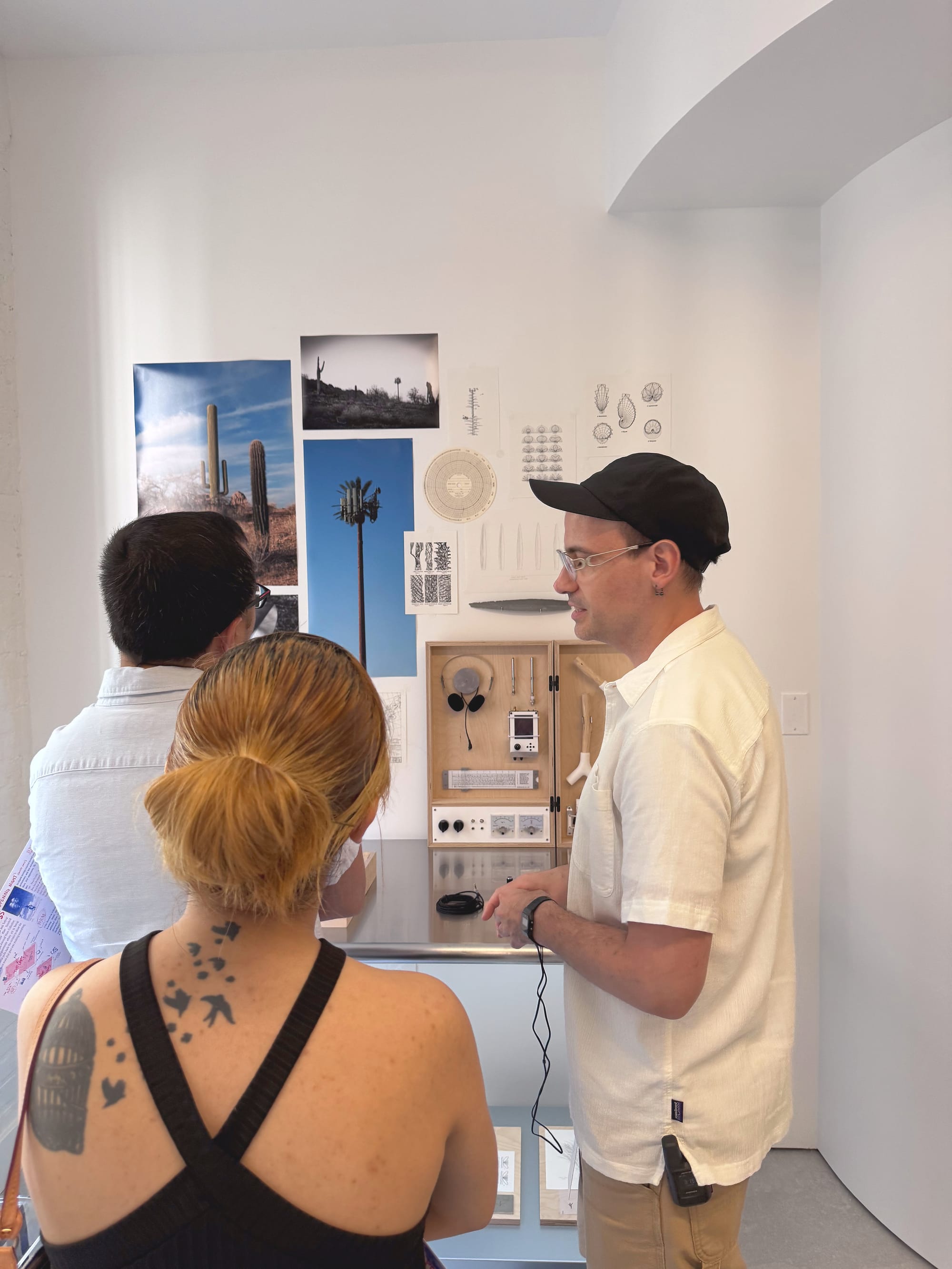 At an art fair booth, a man in a white shirt and black cap talks with two visitors in front of a wall of desert photographs, including a cell tower disguised as a tree, alongside technical diagrams and a wooden case of equipment.