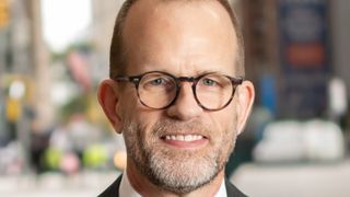 A lawyerly man wearing a suit and glasses is pictured smiling against a city street background 