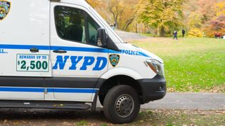 An NYPD police van is pictured on the sidewalk in Central Park in New York City. The grass is green and the leaves are colorful, indicating fall