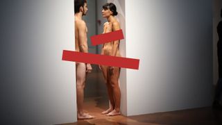 A man and woman stand face each other, nude, in a museum with red censor bars placed over them