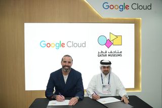 Two men sit in front of logos for Qatar Museums and Google Cloud signing paperwork.