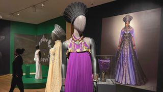 Mannequin dressed in a regal purple Wakanda gown and ornate headpiece on display in a museum gallery, with a large portrait of the character in the background and additional costumes nearby.