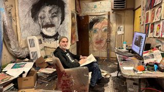 Jon Sarkin sits in an armchair in a messy artist studio