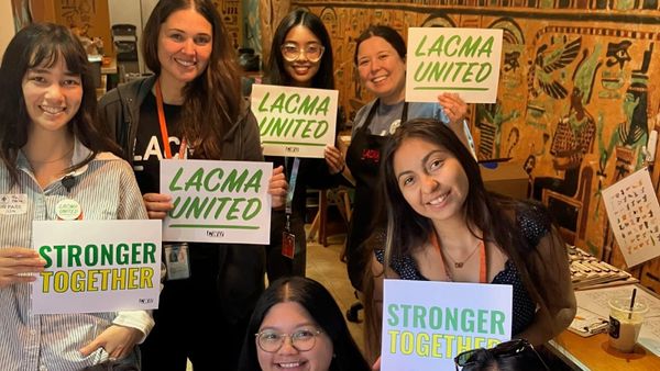 Eight Los Angeles County Museum of Art employees smile and hold signs reading “LACMA United,” “Recognize Our Union,” and “Stronger Together” inside a museum workspace.