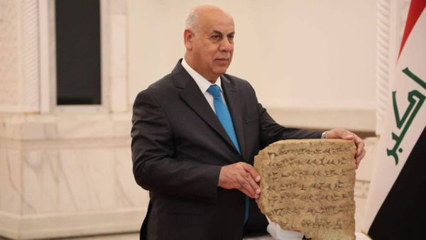 A man in a dark suit and blue tie stands beside an Iraqi flag, holding an ancient clay tablet inscribed with cuneiform script inside a marble hall.