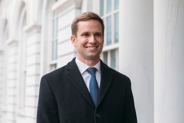 A man with a short haircut, a black suit and blue tie smiles in front of a government building.