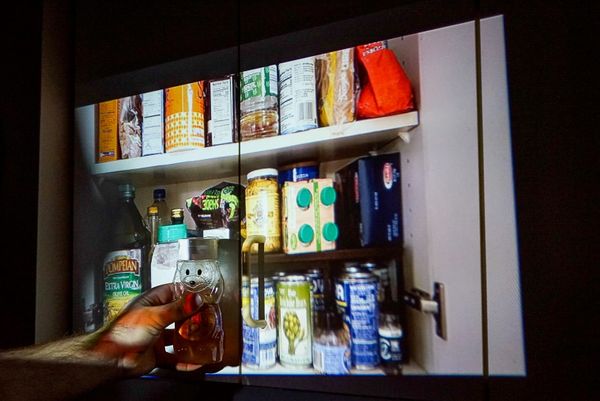 A man's hand is seen holding a honey container up to a kitchen cabinet, with the contents inside the cabinet projected onto its outer doors