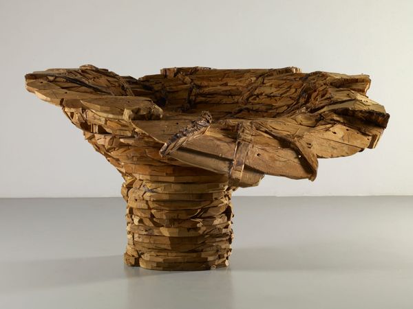 A large, rough-hewn cedar sculpture shaped like a wide, flaring bowl, built from stacked, carved wooden planks with a textured, striated surface.