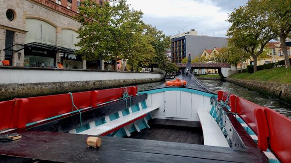 A view from inside a colorful wooden boat moving through a narrow canal lined with trees, buildings, and a small pedestrian bridge ahead.