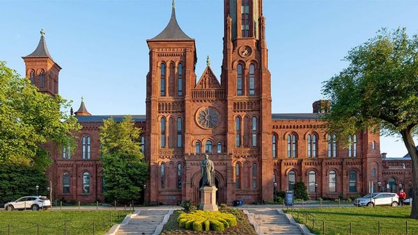 Red-brick Smithsonian Castle on the National Mall, framed by green trees, with a statue at the front entrance under a clear blue sky.