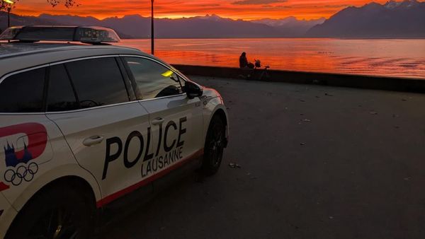 A Lausanne police car parked beside a lakeside promenade at sunset, with vivid orange and red skies reflecting over the water and mountains in the distance