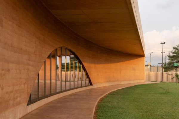 Curved exterior wall of the Museum of West African Art in Benin City, built with warm-toned rammed earth and large arched glass windows reflecting the surrounding landscape.