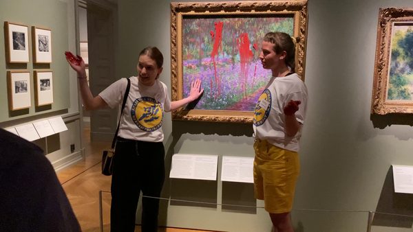 Two climate protesters stand in front of a Monet painting inside a museum gallery, with red paint smeared across the artwork’s protective surface.