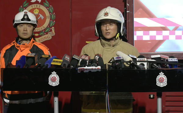 wo Hong Kong Fire Services officers stand at a podium during a nighttime press briefing, surrounded by microphones, with a fire services emblem and emergency vehicle visible behind them.