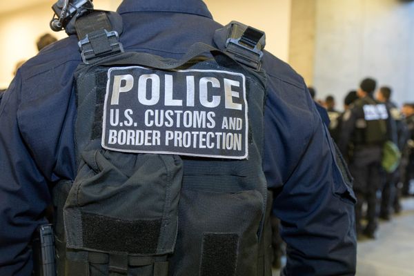 A U.S. Customs and Border Protection officer wearing a tactical vest marked “Police – U.S. Customs and Border Protection,” seen from behind at an indoor enforcement setting.