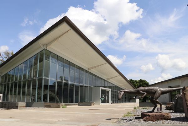 Exterior of the Museum of the Earth in Ithaca, New York, showing the modern glass-fronted building and a dinosaur sculpture outside the entrance.
