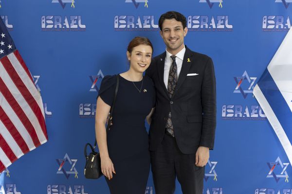Two Israeli Embassy staff members pose for a photo at a diplomatic event, standing in front of an Embassy of Israel backdrop with U.S. and Israeli flags visible.