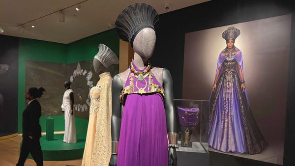 Mannequin dressed in a regal purple Wakanda gown and ornate headpiece on display in a museum gallery, with a large portrait of the character in the background and additional costumes nearby.