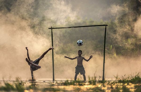 Two boys playing football in golden evening light, one mid-kick between makeshift goalposts