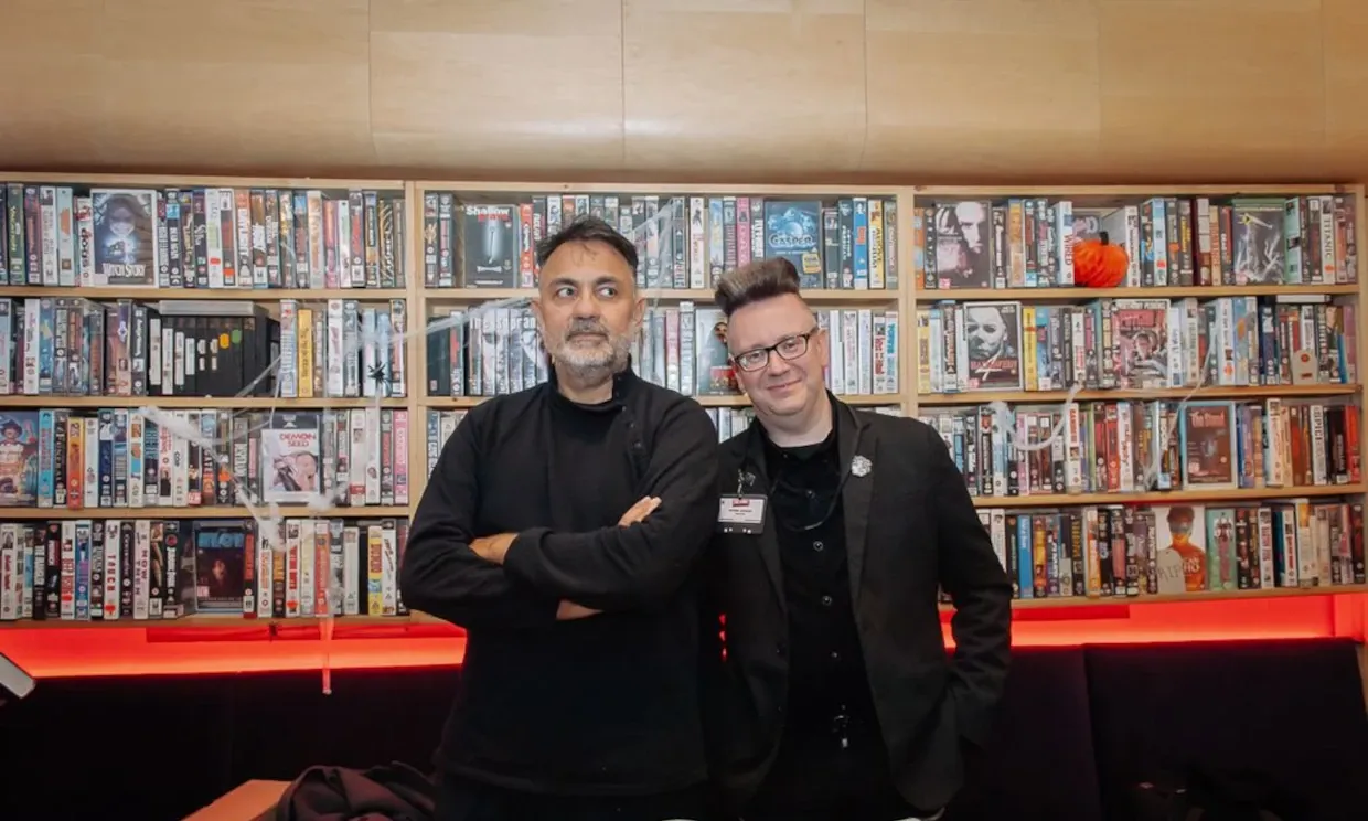 Two men stand in front of shelves of videos. 