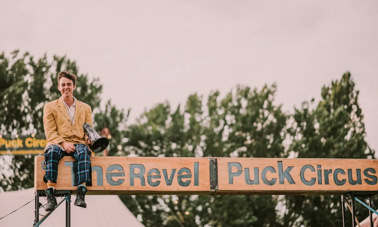  A man holding a megaphone sits atop a sign that reads 'The Revel Puck Circus'. 