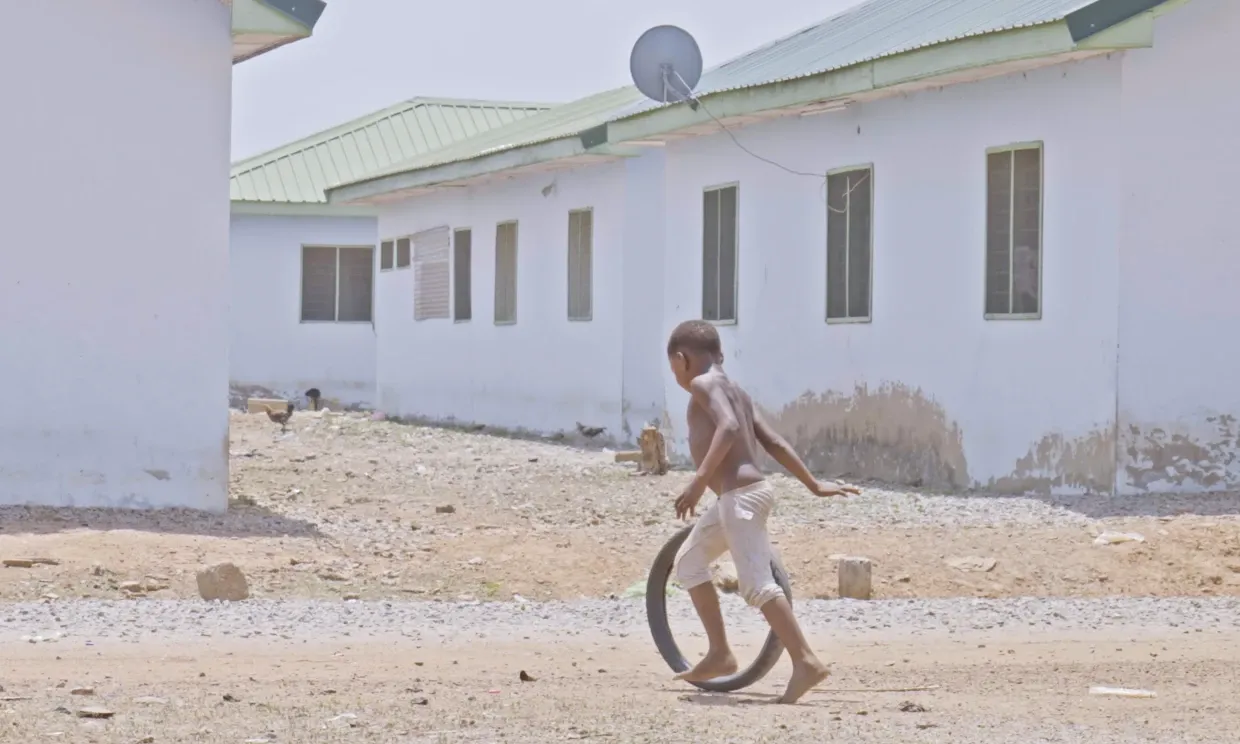 A little boy rolls a tire along a dusty road in front of white houses