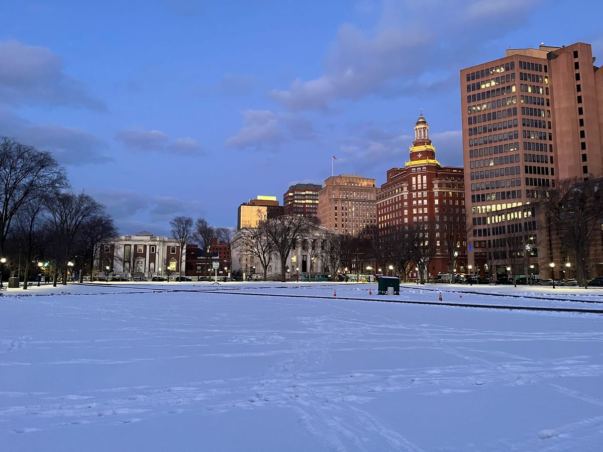 an image of a snow covered public park at dawn. The sky is blue and there are some light clouds. The buildings in the background are reflecting the morning light. Tracks crisscross the field.