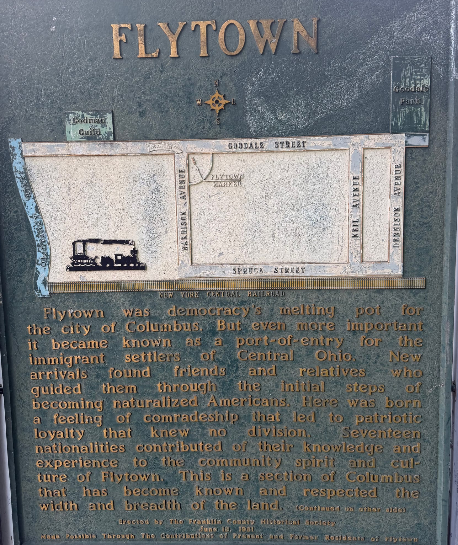 A green plaque with gold lettering telling the story of immigrants that settled the area of Flytown, including seventeen different nationalities.
