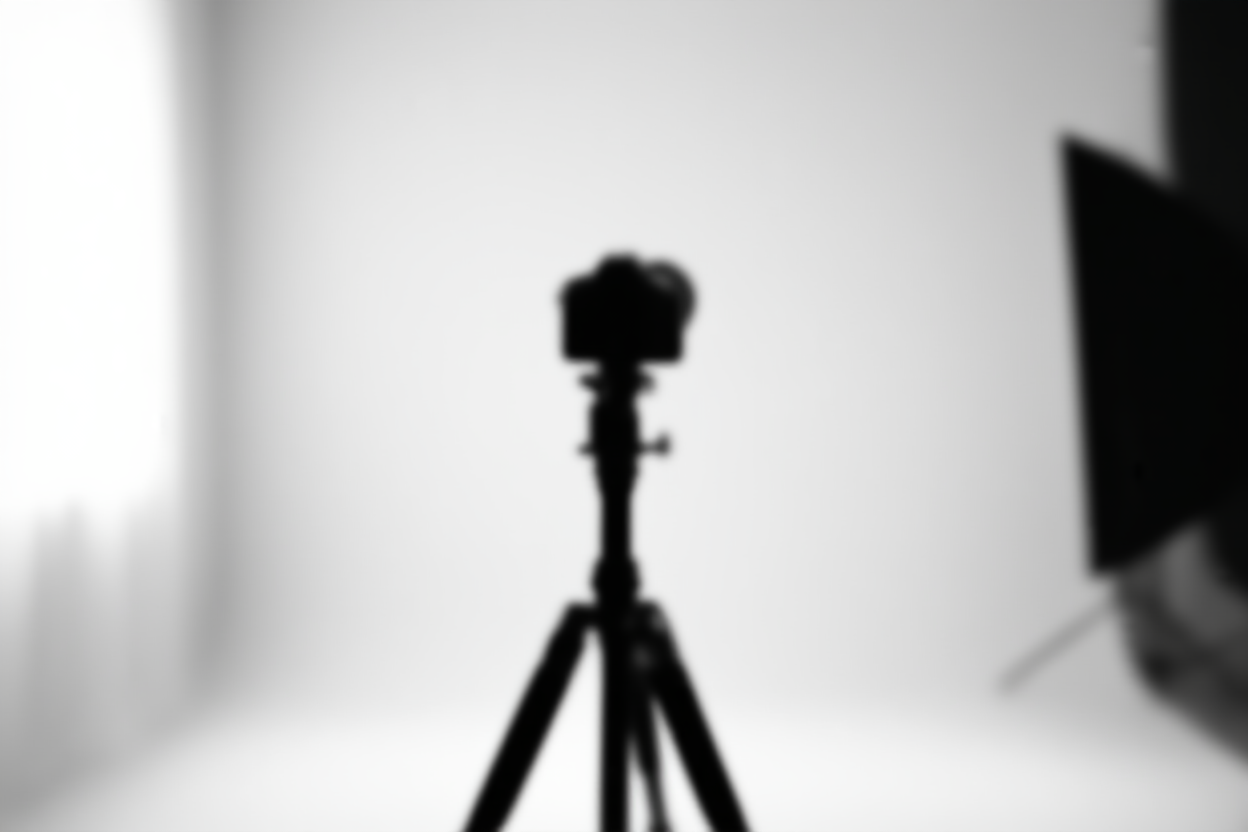 Pengaturan studio yang cerah menampilkan tripod ramping yang memegang kamera mengarah ke latar belakang putih minimalis dengan pencahayaan softbox ligh