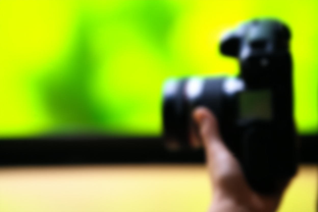 Tay của một nhiếp ảnh gia cầm máy ảnh DSLR với ống kính zoom gắn vào, đứng trong khu vườn đầy nắng với nền xanh lá cây mờ phía sau.