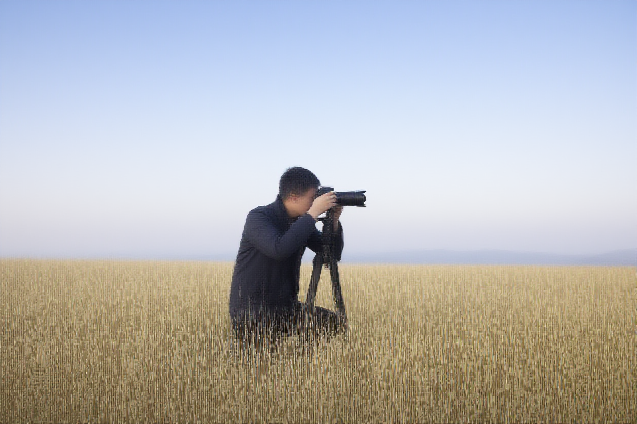 Ein Fotograf kniet im hohen goldenen Gras bei Sonnenaufgang und justiert eine auf einem Stativ montierte DSLR-Kamera, entfernte Berge v