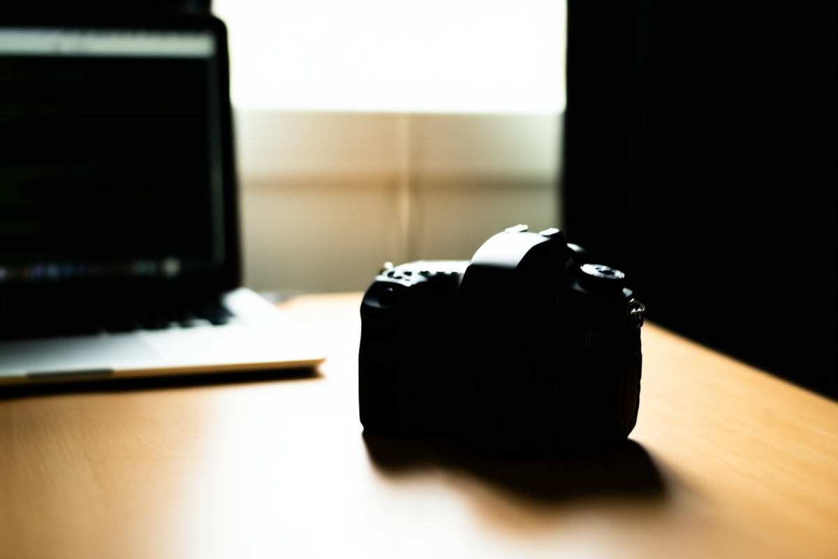 Professionelle DSLR-Kamera auf einem Holzschreibtisch neben einem Laptop, der Code anzeigt, weiches natürliches Fensterlicht