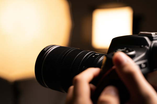 Zbliżenie na dłonie fotografa trzymającego aparat DSLR z założonym kapturem obiektywu, stojącego w jasnym studiu wypełnionym softboxami i parasolami refleksyjnymi przed białym tłem