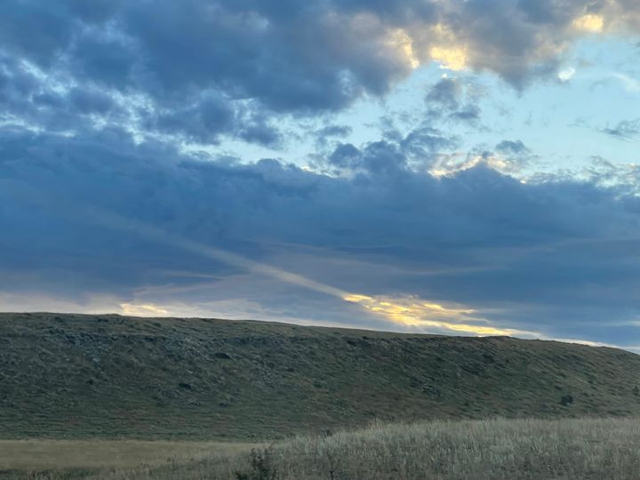 A green grass hill offsets some clouds with a sun shaft in the distance.
