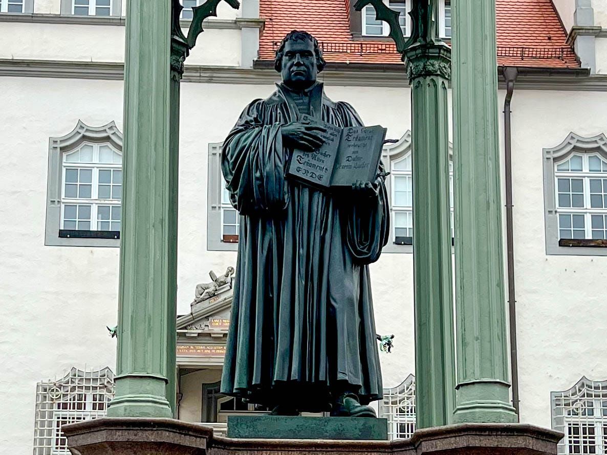 Denkmal Martin Luthers auf einem Sockel und zwischen zwei Säulen auf dem Marktplatz von Wittenberg. Luther hält ein aufgeschlagenes Buch in den Händen.