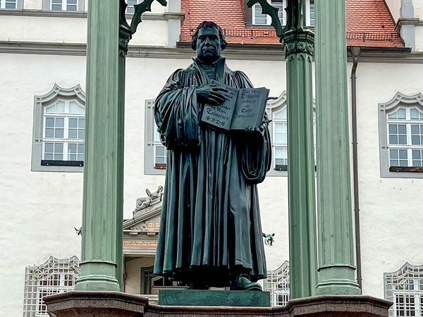 Denkmal Martin Luthers auf einem Sockel und zwischen zwei Säulen auf dem Marktplatz von Wittenberg. Luther hält ein aufgeschlagenes Buch in den Händen.