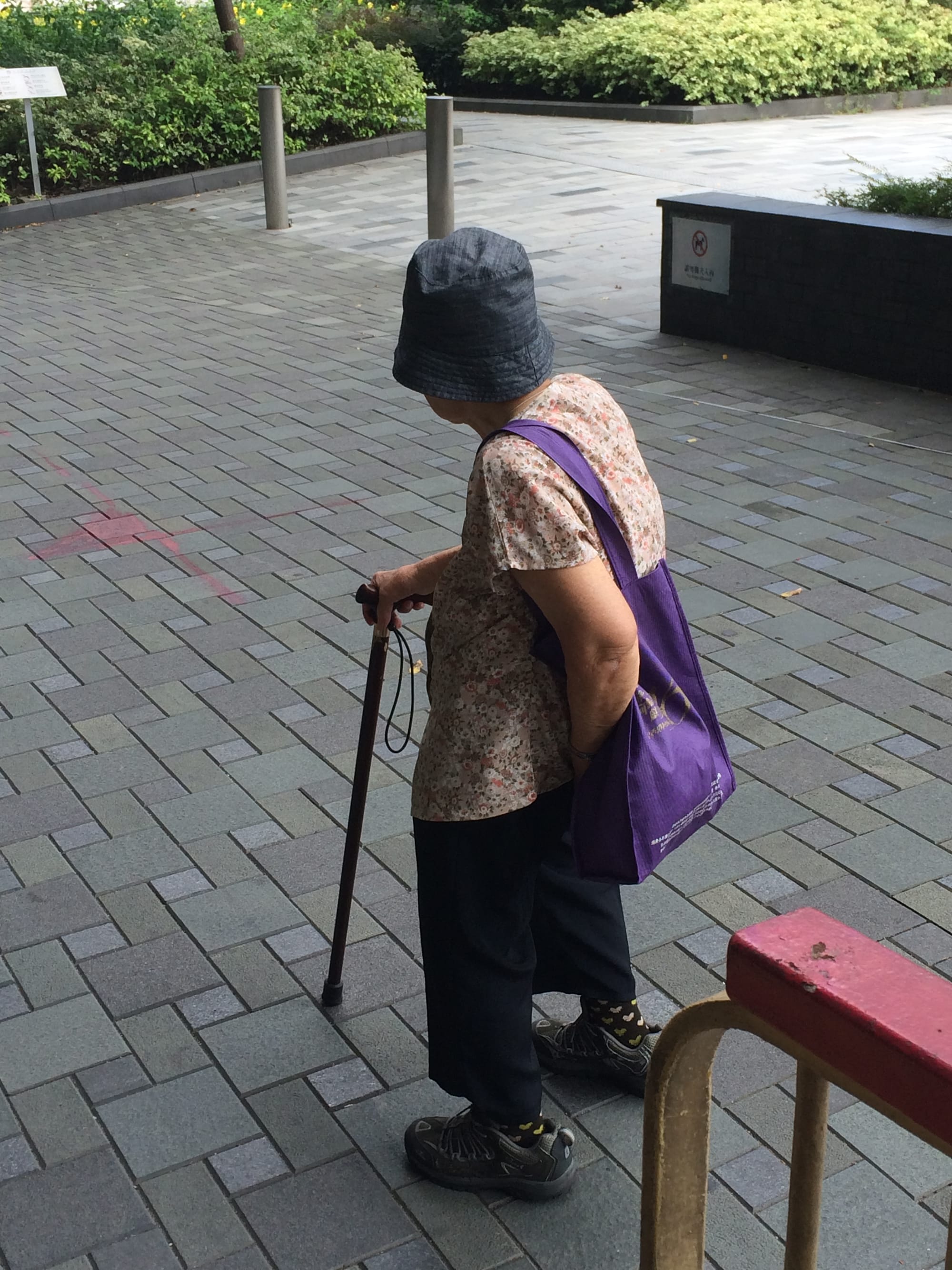 Side view of a granny with a cane. She wears trainers, a denim bucket hat, and carries a purple tote bag.