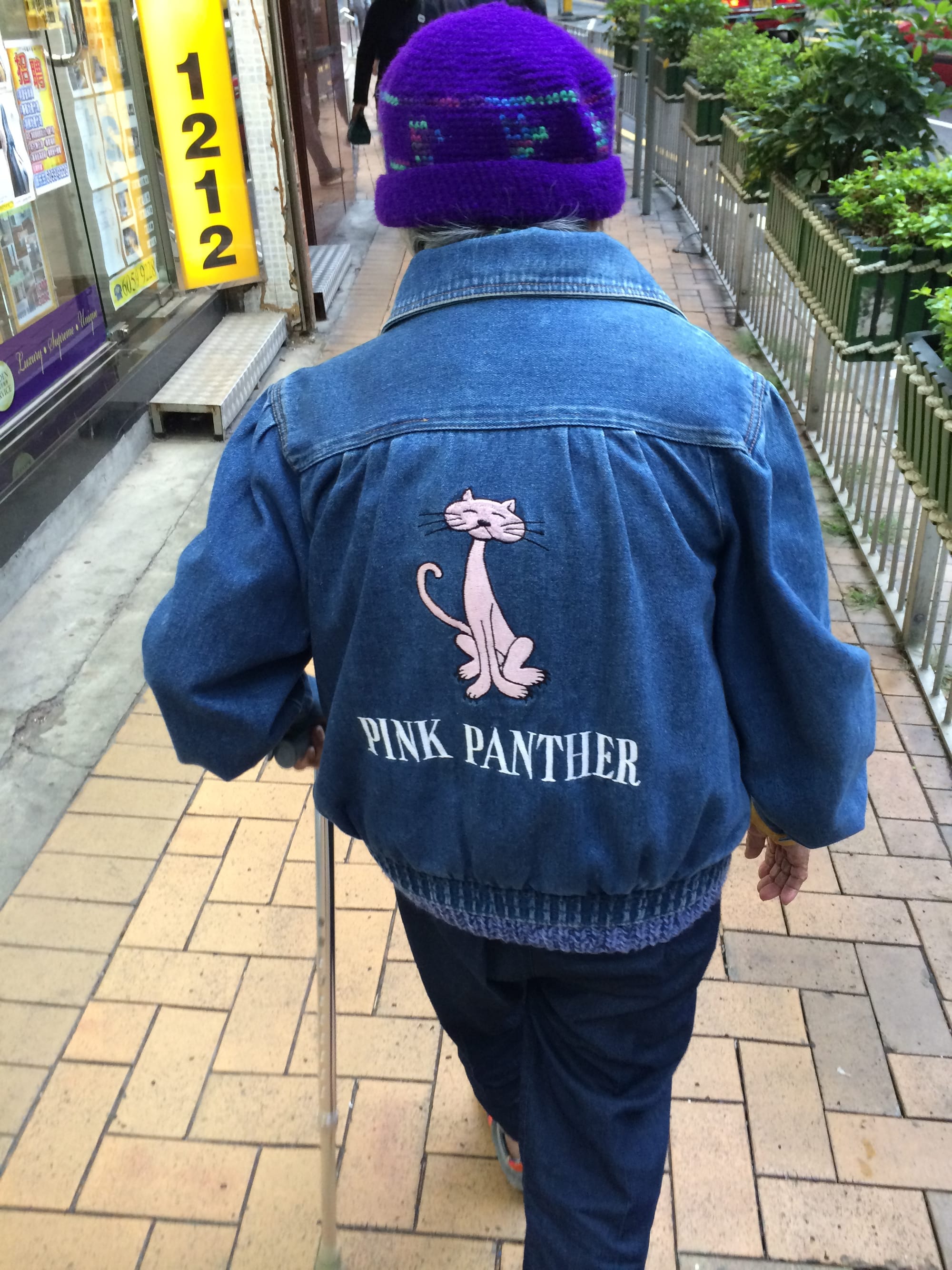 Back view of a granny with a cane, wearing a denim jacket with a pink cat and the words "Pink Panther" embroidered on its back. She also dons a purple beanie.